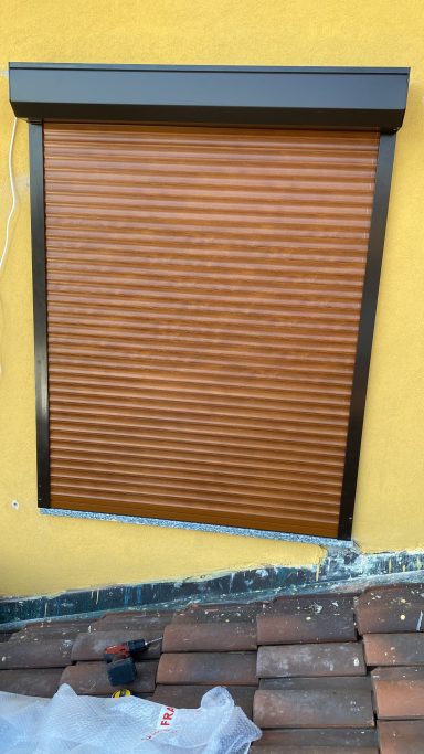 Rolled-up wooden blinds installed on a yellow wall with a tile roof below.